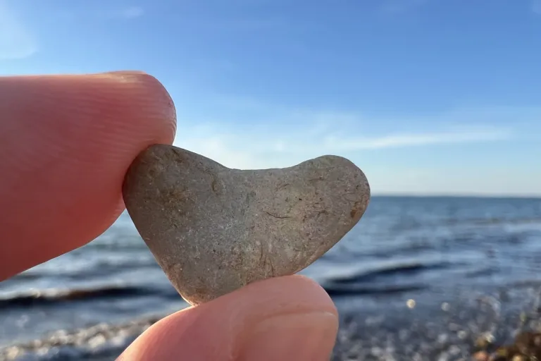 Herzförmiger Stein zwischen zwei Fingern, dahinter das Meer - Heiko Metz