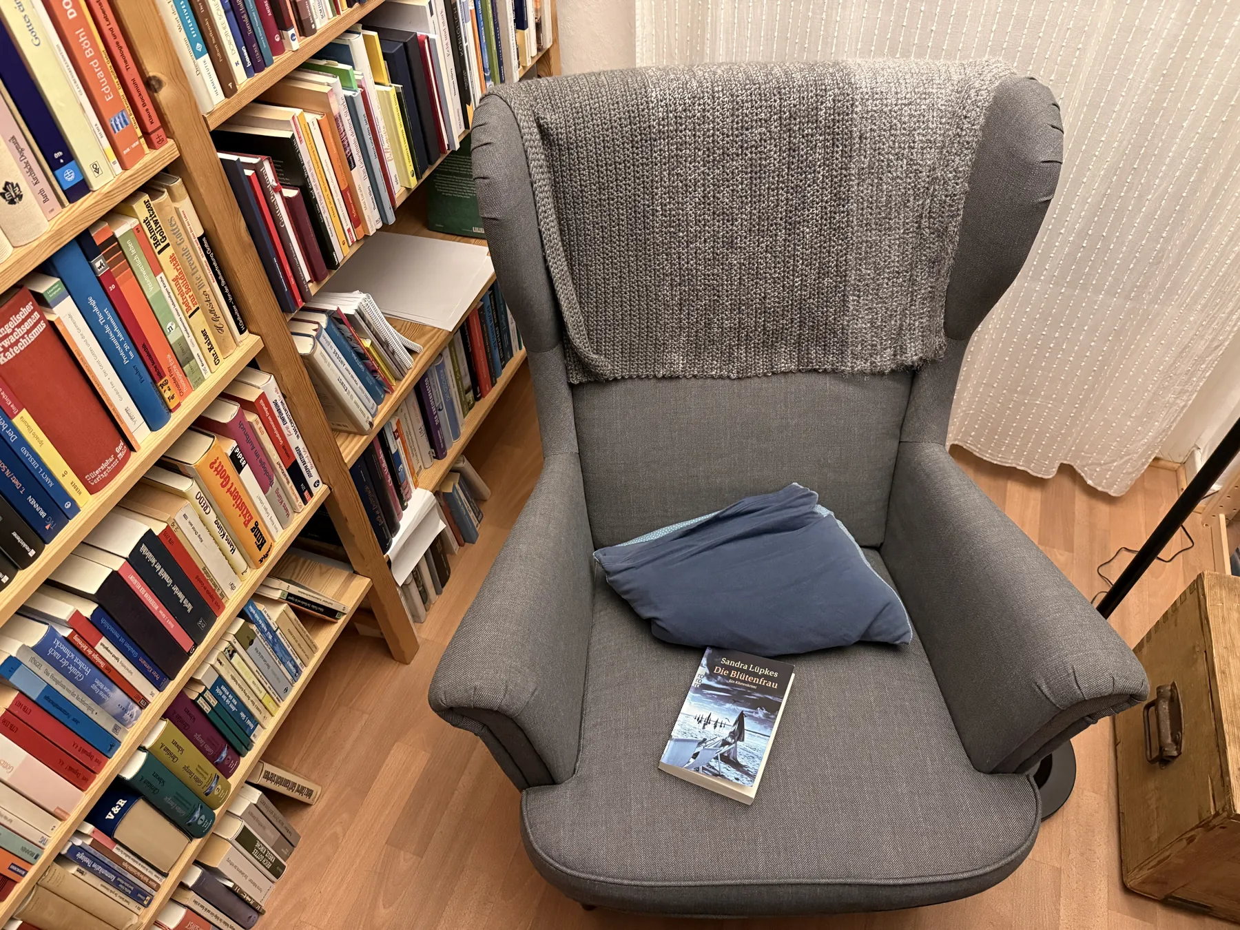 Grauer Ohrensessel mit Decke und einem Buch vor einem vollen Bücherregal - Heiko Metz